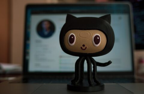 An octocat figurine in front of a blurred out computer screen that is displaying GitHub.